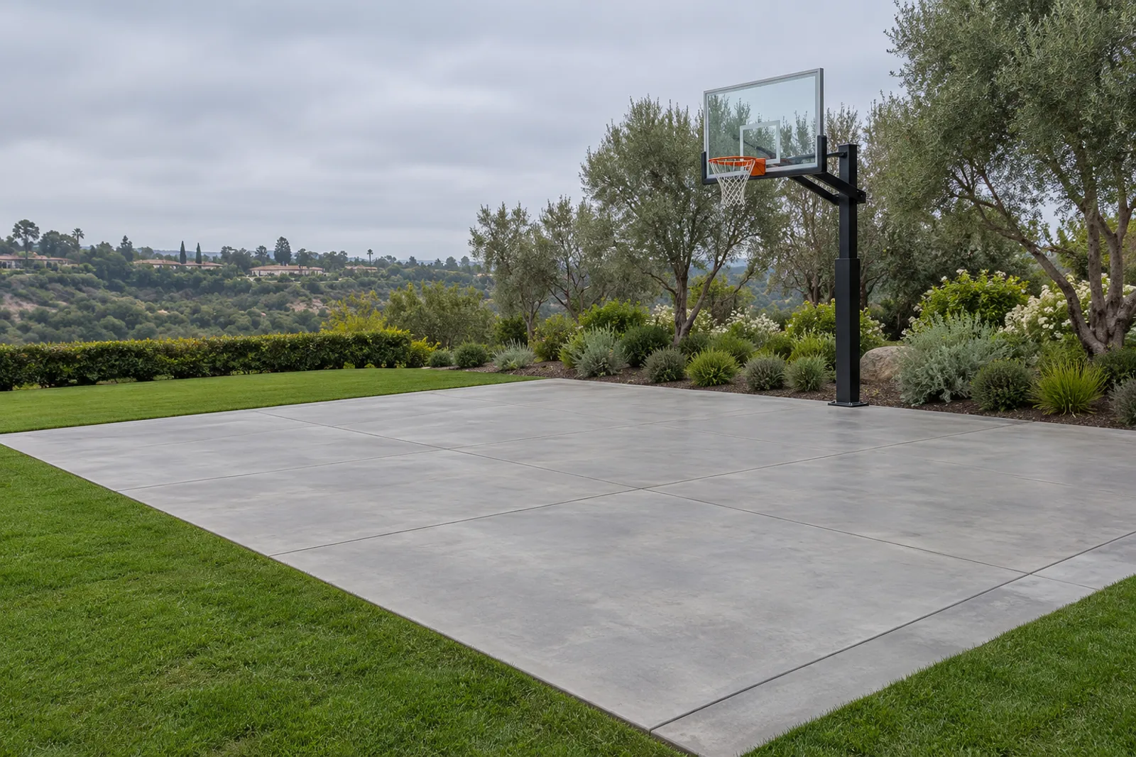 Unmarked steel-troweled concrete half-court slab with saw-cut joints and a basketball hoop on a lawn with olive trees